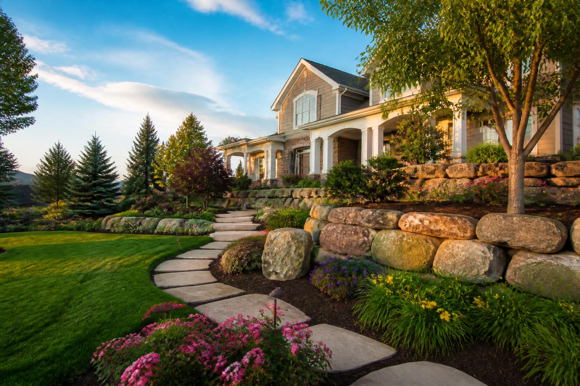 Professionally landscaped property with green lawn, mulch beds, flower beds, boulder retaining wall, and a stone pathway under a blue sky